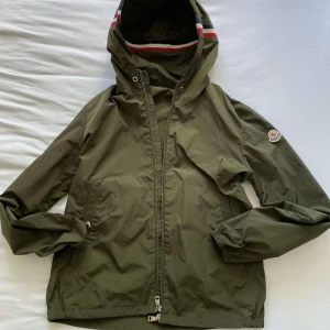 Grön vindjacka från Moncler med huva - Ifall du har ett prisförslag Skriv gärna till mig ifall du är intresserad av en Snygg grön vindjacka från Moncler med huva och dragkedja. Jackan har Moncler-logga på ärmen och röd, vit och blå rand på huvan. Perfekt för blåsiga dagar och enkel att matcha med det mesta.