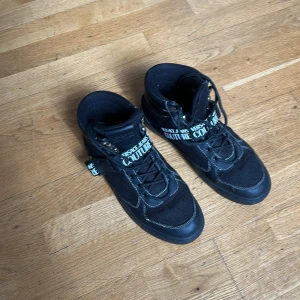 Svarta sneakers från Versace Jeans Couture - Säljer ett par svarta sneakers från Versace Jeans Couture med vita logodetaljer på sidan och snörning. Skorna har hög modell, rund tå och är tillverkade i en kombination av syntet och textil. Perfekta för dig som vill sticka ut med en exklusiv streetstyle.