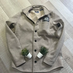 Beige overshirt från Jack & Jones - Säljer en stilren beige skjorta från Jack & Jones med svarta knappar och en bröstficka. Skjortan har klassisk krage och är tillverkad i ett mjukt material som passar perfekt till vardags. Enkel och tidlös design. Den är endast testad så nyskick! Pris går o diskutera!