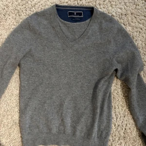Grå v-ringad tröja från Hansen & Jacob - Säljer en stilren grå v-ringad tröja från Hansen & Jacob i 100% lammull. Perfekt för lager-på-lager och passar till många olika outfits. Klassisk modell med ribbade muddar vid ärmslut och nederkant.