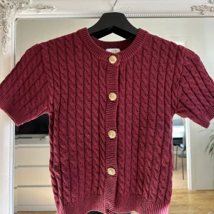 Vinröd kabelstickad kofta med kort ärm - Säljer en vinröd kabelstickad kofta med korta ärmar och dekorativa guldknappar framtill. Perfekt för lager-på-lager och snygg till både jeans och kjol.