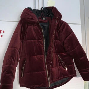 Vinröd pufferjacka med huva från Only - Mysig vinröd pufferjacka från Only med huva och guldfärgade dragkedjor. Jackan är vadderad och har två fickor framtill. Perfekt för kyligare dagar och ger en snygg look till vardags.