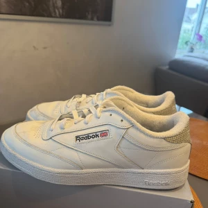 Vita Reebok sneakers med rutigt mönster - Säljer ett par klassiska vita Reebok sneakers med rutigt mönster på hälen och Reebok-logga på sidan. Skorna har snörning, rund tå och platt sula. Perfekta för en stilren och sportig look.