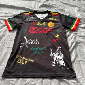 Ajax x Bob Marley special edition fotbollströja - Unik svart Ajax fotbollströja i samarbete med Bob Marley, med färgglada detaljer i rött, gult och grönt på ärmarna. Tryck med Ziggo-logga, Ajax-märke, Bob Marley-motiv och inspirerande texter. Kortärmad och tillverkad i polyester.