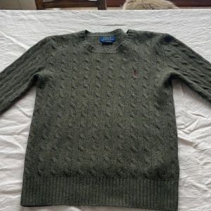 Olivgrön stickad tröja från Polo Ralph Lauren - Säljer en olivgrön stickad tröja från Polo Ralph Lauren med klassiskt kabelstickat mönster och rund halsringning. Tröjan har lång ärm och liten broderad logga på bröstet. Perfekt för lager-på-lager-stil. Passar dig ifrån 158-170