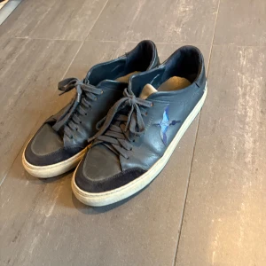 Svarta sneakers med broderad fågel - Snygga svarta sneakers från Axel Arigato med broderad blå fågel på sidan. Skorna har sett bättre dagar men har fortfarande mycket kvar att ge och är ferfekta att använda som beaters.