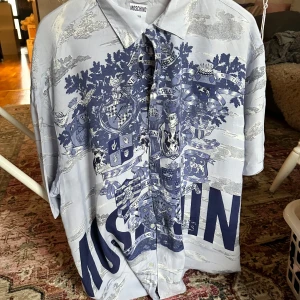 Äkta retro 90-tals Moschino skjorta – unisex, blå m. tryck, oversized - Äkta 90-tals Moschino! Unisex kortärmad skjorta i blå toner med konstnärligt tryck – ikonisk och oversized passform. Från Moschino Jeans, vintage och i mycket fint skick. Cool, unik och full av personlighet. Storlek XS men passar även M/L beroende på stil. Ett riktigt kap för modeälskaren!
