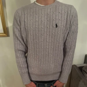 Ljusgrå kabelstickad tröja från Ralph Lauren - Säljer en ljusgrå kabelstickad tröja från Polo Ralph Lauren med klassisk broderad logga på bröstet. Tröjan har rund halsringning och långa ärmar. Perfekt för lager-på-lager och stilrena outfits. Modellen i bilden är 180cm 65kg. Nypris för tröjan är ca 2400kr.
