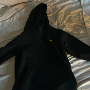 Svart hoodie från Lyle & Scott - Säljer en svart hoodie från Lyle & Scott med klassisk gul logga på bröstet. Tröjan har huva och en stor magficka framtill. Perfekt för dig som gillar stilrena och bekväma plagg.