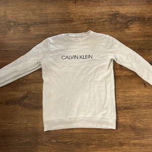 Vit sweatshirt från Calvin Klein - Säljer en stilren vit sweatshirt från Calvin Klein med svart logotyptryck på bröstet och liten CK-detalj på ärmen. Tröjan har rund halsringning och långa ärmar. Perfekt för en clean och klassisk look.