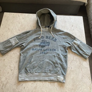 Grå hoodie från Polo Ralph Lauren - Säljer en grå hoodie från Polo Ralph Lauren med trycket 'Polo Bear Ralph Lauren 1967' och en björn på framsidan. Tröjan har huva med dragsko och en stor magficka. Perfekt för en avslappnad stil. Nypris:ca 3000kr