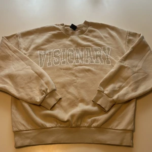 Beige sweatshirt med tryck från Gina Tricot - Säljer en beige sweatshirt från Gina Tricot med texten 'VISIONARY' tryckt framtill. Tröjan har rund hals, långa ärmar och ribbade muddar vid ärmslut och nederkant. Perfekt för en avslappnad och trendig stil.