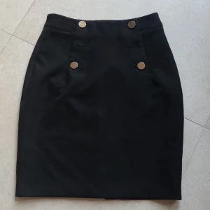 Svart pennkjol med guldknappar från H&M - Klassisk svart pennkjol från H&M med fyra dekorativa guldknappar framtill. Kjolen har hög midja och dragkedja bak. Perfekt till en snygg vardagslook eller när du vill känna dig extra stilig.