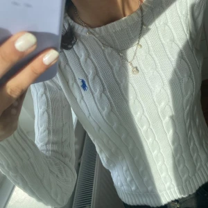 Vit kabelstickad tröja från Polo Ralph Lauren - Säljer en klassisk vit kabelstickad tröja från Polo Ralph Lauren med blå broderad logga på bröstet. Tröjan har rund halsringning, långa ärmar och ribbade muddar vid ärmslut och nederkant. Perfekt för en stilren och tidlös look. Har inte använt den så mycket💕🌸