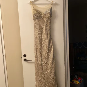 Glittrig långklänning med pärldetaljer som är en festlig klänning - Säljer en glittrig långklänning i champagnefärg med smala axelband och v-ringning. Klänningen har pärlor och paljetter upptill samt ett glittrigt mönster över hela tyget. Perfekt för bal eller fest! Materialet känns lyxigt och skimrande.