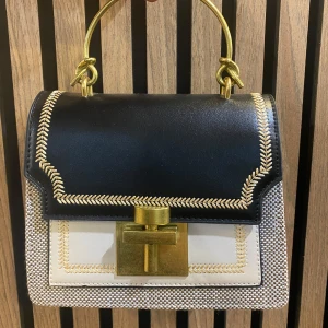 Svart och vit handväska med gulddetaljer - Snygg handväska med svart lock och vit framsida, dekorerad med guldfärgade sömmar och ett stort guldfärgat spänne. Väskan har ett stadigt handtag i metall och en elegant, boxig form. Perfekt accessoar för att lyfta din outfit.