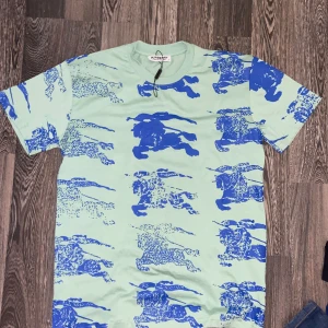 Ljusgrön t-shirt från Burberry - Cool ljusgrön t-shirt från Burberry med unikt blått tryck av riddare och hästar över hela tröjan. Klassisk rund hals och korta ärmar. Perfekt för dig som vill sticka ut med en statement-piece från ett ikoniskt märke.