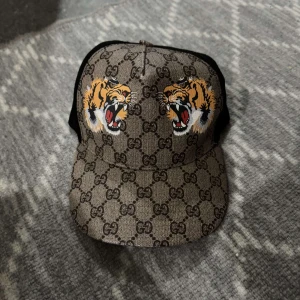 Gucci keps  - Säljer en brun keps från Gucci med klassiskt GG-mönster och broderade tigrar framtill. Kepsen har svart mesh på baksidan och justerbart spänne. Perfekt för dig som vill sticka ut med en exklusiv accessoar.