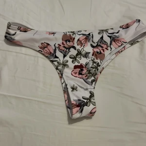 Vit blommig bikiniunderdel - Säljer en vit bikiniunderdel med blommigt mönster i rosa och grönt. Modellen har en klassisk tanga-form och är perfekt för stranden eller poolen. Tyg i syntetmaterial som torkar snabbt. 