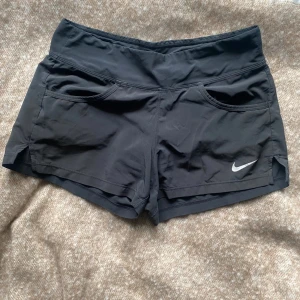 Svarta träningsshorts från Nike - Svarta träningsshorts från Nike med två framfickor och vit logga på vänster ben. Shortsen har en normal passform och är tillverkade i ett lätt syntetmaterial som passar perfekt för träning.