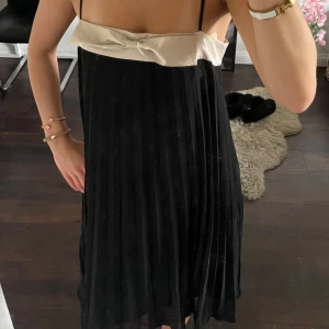 Svart och beige plisserad klänning med rosett - Söt klänning med svart plisserad kjol och beige överdel. Smala axelband och en liten rosett över bröstet 