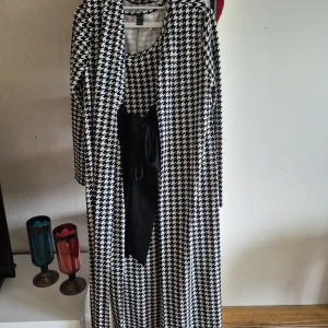 Svartvit långklänning med kavaj och knytband - Säljer en svartvit långklänning med klassiskt hundtandsmönster. Klänningen är ärmlös och har ett svart knytband i midjan. Till klänningen hör en matchande långärmad kavaj i samma mönster. Perfekt för dig som gillar retrostil!