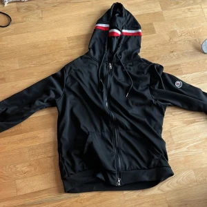 Svart hoodie med dragkedja från Moncler - Svart hoodie från Moncler med dragkedja framtill och huva. Jackan har röda och vita ränder på huvan samt Moncler-logga på ärmen. Praktiska fickor framtill och ribbade muddar vid ärmslut och nederkant. Stl M