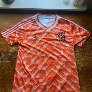 Holland retro fotbollströja Adidas Gullit 10 - Säljer en ikonisk orange Holland retro fotbollströja från Adidas med klassiskt geometriskt mönster, svarta detaljer och tryck KNVB samt nummer 10 och Gullit på ryggen. V-ringad krage och vita ränder på axlarna.
