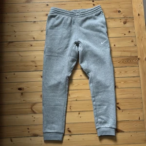 Grå mjukisbyxor från Nike strl M - Snygga grå mjukisbyxor från Nike med standard fit och avsmalnande ben. Byxorna har resår i midjan, sidofickor och en bakficka. Perfekta för en avslappnad stil och har en liten vit Nike-logga på benet.