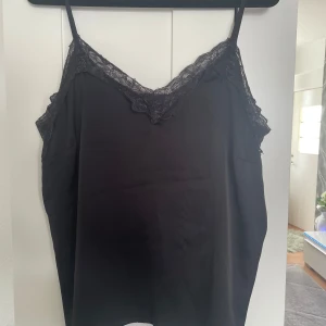 Svart linnetopp med spets - Säljer en svart linnetopp med tunna axelband och v-ringning dekorerad med spets. Perfekt att styla till olika outfits för en trendig look.