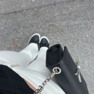 Svart kuvertväska med silverkedja - Säljer en stilren svart kuvertväska med silverkedja och dekorativt hänglås framtill. Väskan har en elegant och enkel design, perfekt att bära över axeln eller i handen. Passar till många olika outfits.