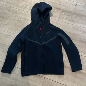 Svart hoodiejacka från Nike - Svart hoodiejacka från Nike med dragkedja framtill och huva. Jackan har en sportig look med diskreta detaljer och logga på bröstet. Perfekt för träning eller chill. Snygg och enkel design som passar till mycket.