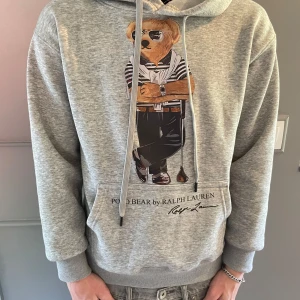 Grå Ralph Lauren Hoodie  - Tjena, Säljer nu en grå hoodie från Polo Ralph Lauren med ikoniskt björntryck på bröstet. Tröjan är i utmärkt skick och är perfekt för dig som vill ha en skön stilren tröja för ett dunder pris. 