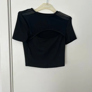 Svart croppad topp med meshdetaljer - Säljer en svart croppad topp med korta ärmar och meshdetaljer på axlarna. Toppen har en snygg cut-out detalj framtill som ger en cool look. Perfekt att styla till jeans eller kjol.