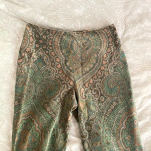 70s vintage byxor från Allan Walker  - Unika byxor från Allan Waller med paisleyinspirerat mönster i grönt, beige och brunt. Byxorna har en normal passform och är tillverkade i ett mjukt material som känns lyxigt. Perfekta för dig som vill sticka ut med en färgstark och retro look.
