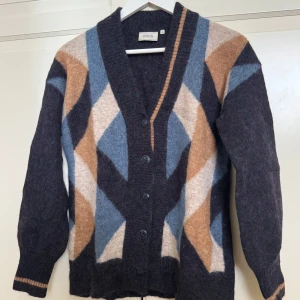 Mönstrad kofta från Boomerang - Säljer en supermjuk och mysig kofta från Boomerang i storlek S. Koftan har ett grafiskt mönster i blått, beige och brunt, med djup v-ringning och knappar framtill. Tillverkad i en stickad ullblandning, perfekt för lager-på-lager.