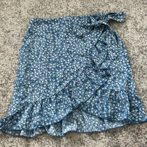 Blå blommig omlottkjol med volang - Supersöt blå omlottkjol med vitt blommönster och volangdetalj längs kanten. Oppnas med en kedja på baksidan. Kjolen knyts i midjan och har en luftig känsla, perfekt för varmare dagar. Passar dig som gillar en feminin och perfekt för sommar❤️