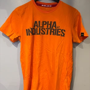 Orange t-shirt från Alpha Industries - Snygg orange t-shirt från Alpha Industries med stor svart logga på bröstet. Klassisk rund halsringning och korta ärmar. Perfekt för dig som vill sticka ut med färg och stil.
