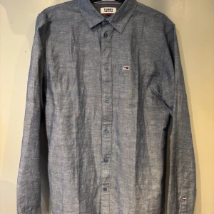 Blå linneskjorta från Tommy Jeans - Säljer en stilren blå långärmad skjorta från Tommy Jeans i regular fit. Skjortan är tillverkad i linne och har klassisk krage, knappar framtill och diskret logga på bröstet. Perfekt för en avslappnad men snygg look.