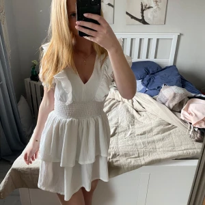 Vit volangklänning med v-ringning - Säljer en söt vit klänning med v-ringning och volangdetaljer. Klänningen har korta puffärmar och smock i midjan för en bekväm passform. Perfekt för sommarens alla tillfällen!