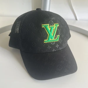 Svart keps från Louis Vuitton med grön logga - Svart keps med mesh baktill och broderad LV-logga i grönt och gult framtill. Bak på kepsen står det 'Louis Vuitton' broderat i grönt. Justerbart spänne i mässing på baksidan. Klassisk böjd skärm och diskret mönster i tyget.