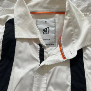Vit skjorta med svarta detaljer från Mouli - Säljer en vit skjorta från Mouli med svarta vertikala ränder framtill och en orange detalj på insidan av kragen. Skjortan har klassisk krage och knäppning framtill. Perfekt för dig som vill sticka ut lite extra.