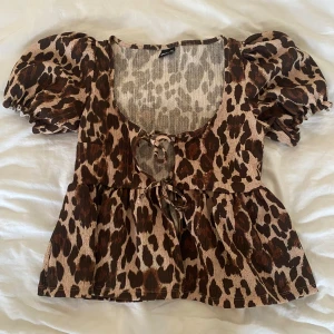Leopardmönstrad blus med puffärmar  - Säljer en leopardmönstrad blus från Gina Tricot med korta puffärmar och två knytningar framtill. Blusen har en luftig passform och är tillverkad i ett lätt, strukturerat tyg. Perfekt inför sommaren. Toppen är aldrig använd!!