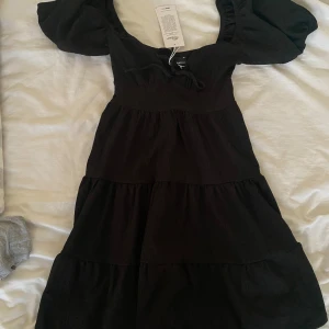 Svart klänning med puffärmar från Gina Tricot - Supersöt svart klänning från Gina Tricot med puffiga kortärmar och knytning framtill. Klänningen har en smickrande v-ringning och volanger i kjolen som ger en fin siluett. Perfekt för dig som gillar trendiga och feminina plagg. Aldrig använd och taggen sitter kvar