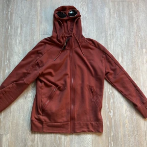 Vinröd ziphoodie från C.P. Company - Säljer en unik vinröd ziphoodie från C.P. Company med inbyggda glasögon i huvan. Jackan har dragkedja framtill, två snedställda fickor med dragkedja och justerbar huva med snörning. Perfekt för dig som vill sticka ut med en streetwear-look.
