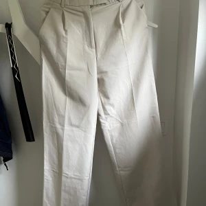 Beiga kostymbyxor från Zara - Snygga beiga kostymbyxor från Zara med hög midja och pressveck. Byxorna har raka ben och diskreta fickor, perfekta för en stilren look. Tillverkade i ett mjukt bomullsmaterial som känns bekvämt hela dagen.