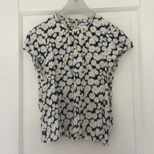 Blommig blus från Holly & Whyte - Säljer en söt blommig blus från Holly & Whyte by Lindex i storlek XS. Blusen har ett mönster av vita och gula prästkragar på mörkblå botten och korta ärmar. Perfekt för vår och sommar!