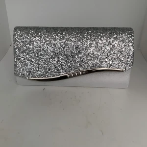 Silverfärgad glittrig kuvertväska med kedja - Snygg silverfärgad kuvertväska med glittrig framsida och elegant metall-detalj. Väskan har en avtagbar silverkedja så den kan bäras både i handen eller över axeln. Perfekt för att lyfta din outfit till nästa nivå.