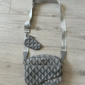 Dior väska - Säljer en snygg grå axelväska från Dior med geometriskt mönster och bred axelrem med logga. Väskan har flera fack och avtagbara delar, perfekt för att hålla ordning på dina grejer. Kommer med tillhörande dustbag. Helt ny och är perfekt inför sommaren, hör av er vid minsta lilla fundering!!👍🏼☺️