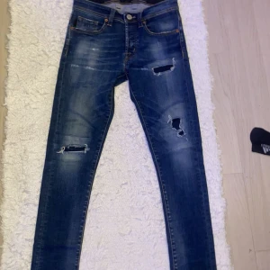Blå slitna jeans från MCMLXVII - Säljer ett par blå jeans från MCMLXVII i storlek w29. Jeansen har flera slitningar och hål på framsidan för en trendig look. Klassisk femficksmodell med smal passform och orange sömmar. Perfekta för dig som gillar en edgy stil. Ny pris 5000kr!